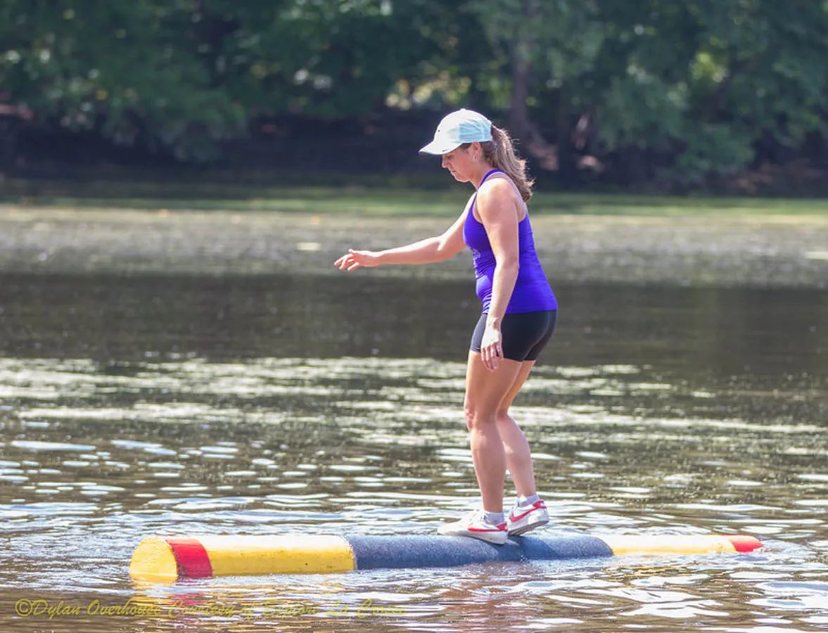 woman in purple tank top and black shorts on top of log in water log rolling
