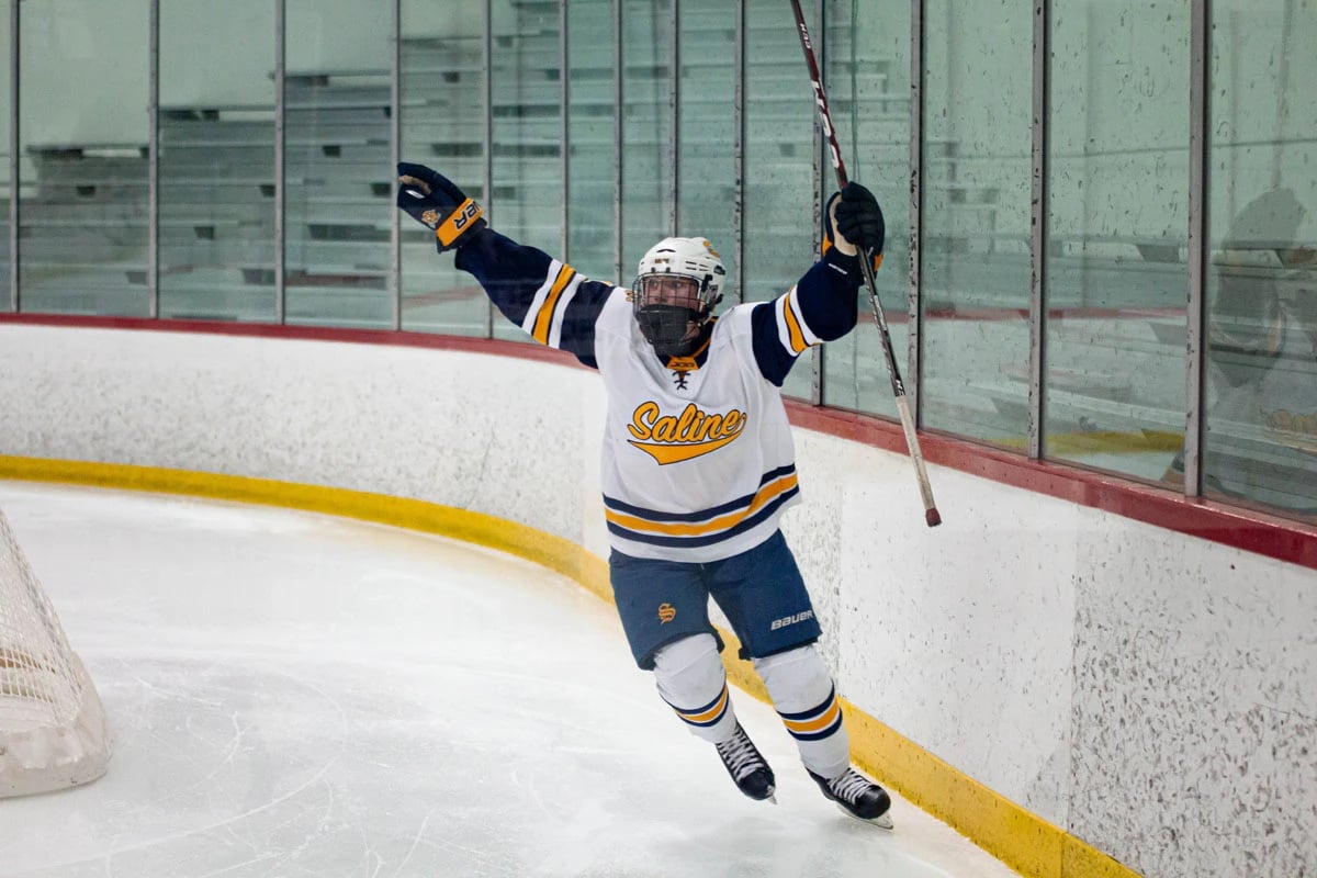Hockey player on the ice with both hands raised in air holding hockey stick up in one hand.
