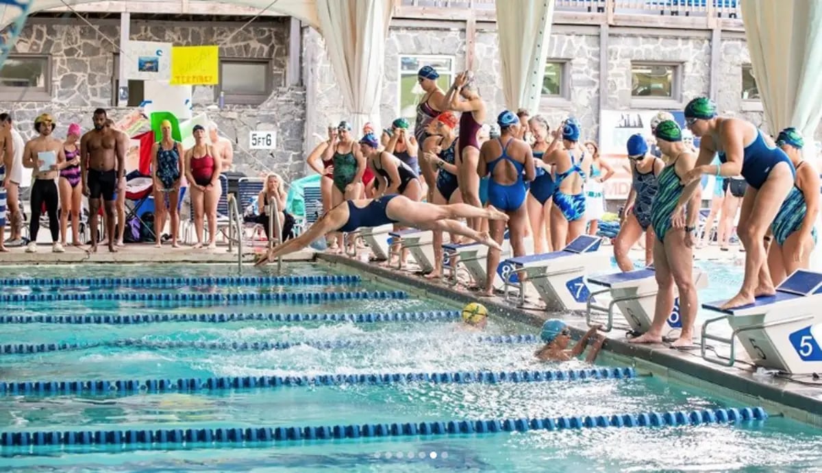 approximately 20 swimmers around a pool at a swim meet, with two swimmers in the pool and one diving in
