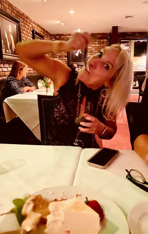 Woman with a spoon in her mouth, wine in her hand at a restaurant and cheesecake in front of her showing excess calorie consumption
