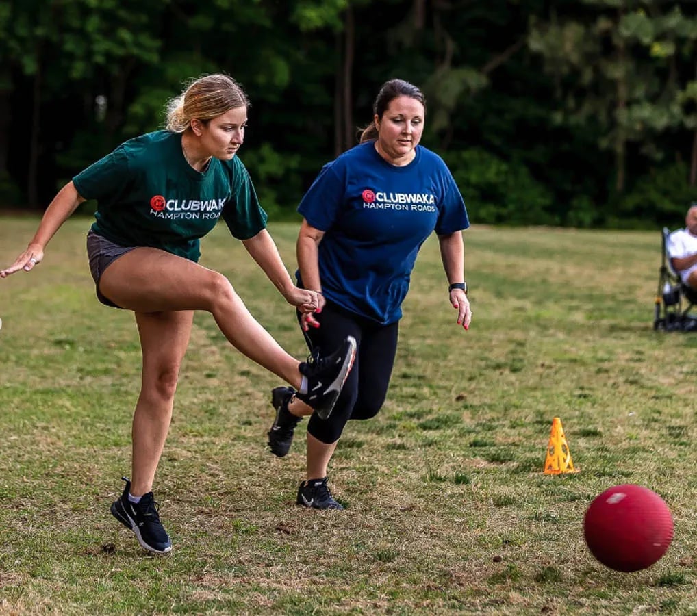 blonde woman kicking a kickball while a brunette woman stands nearby