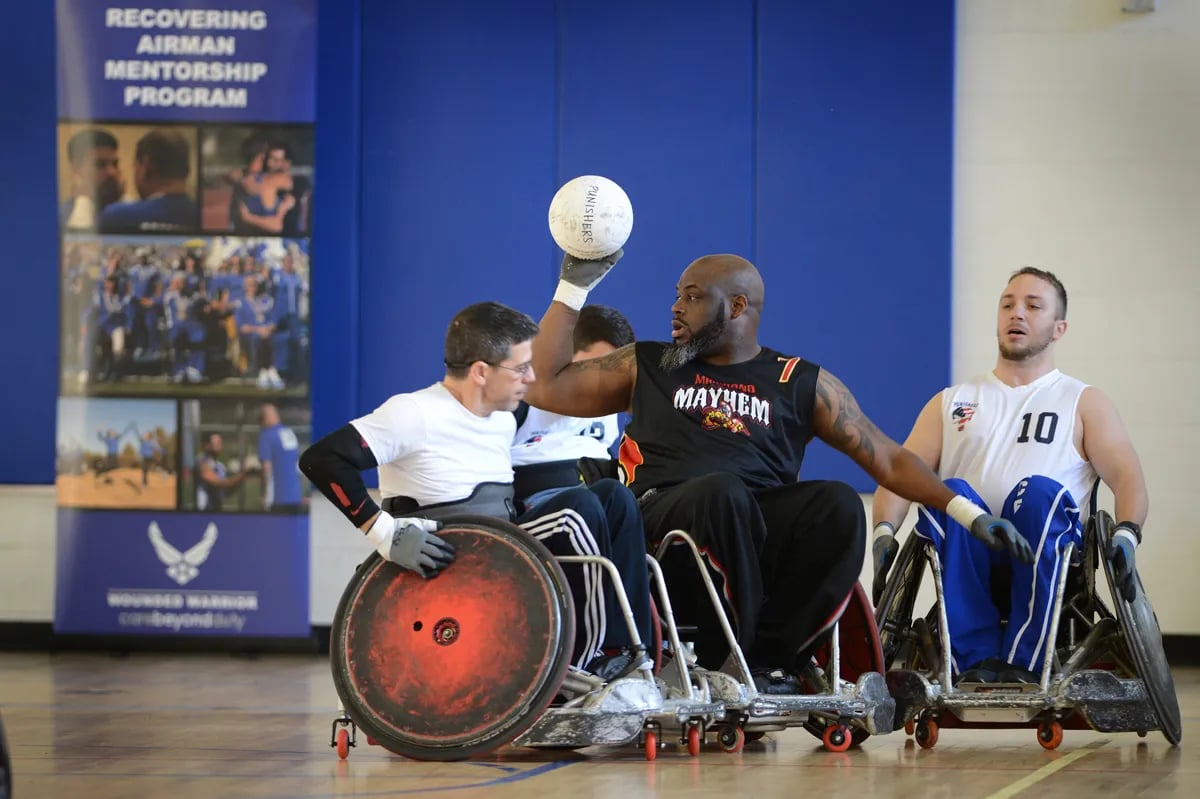 male wheelchair rugby players with one player in black holding the ball in air while three opposing players look on