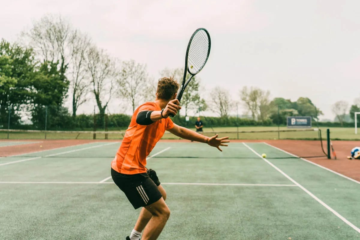 male tennis player with orange shirt and racket raised to hit ball below him across the net to opponent
