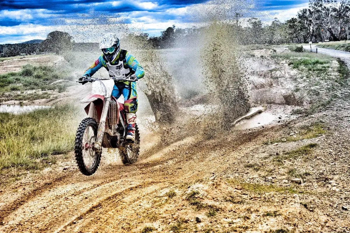 motocross rider in teal blue and lime green top and pants riding through dirt with dirt flying up behind them