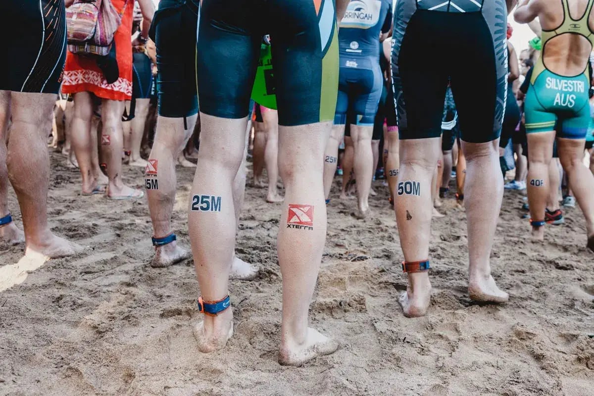 waist down view of triathletes with shorty wetsuits standing in sand and legs marked with numbers