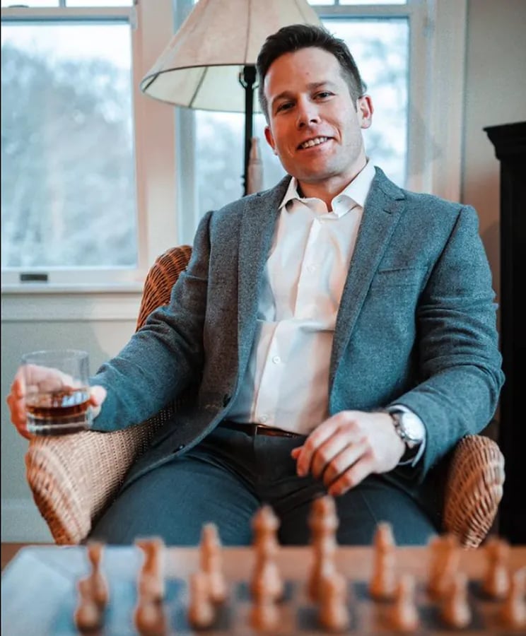 Alex sitting at a chess board with a glass of whiskey in a suit