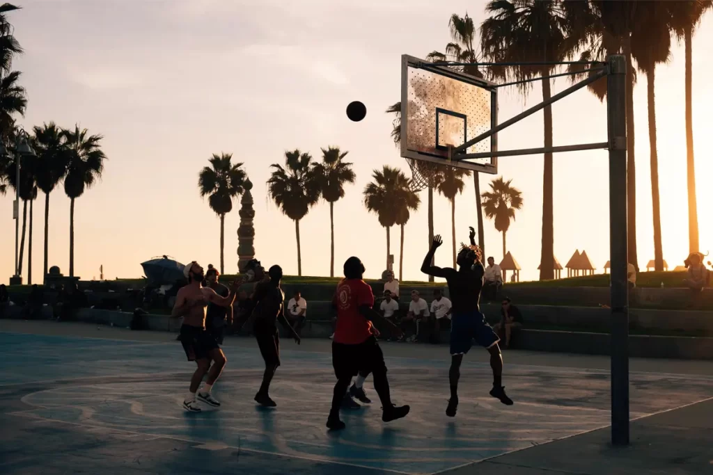 Men playing a pickup basketball outside at sunset with palm trees in the background
