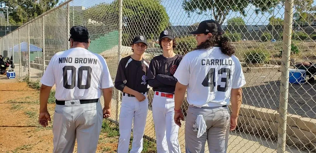 Three generations of Carrillo baseball boy in baseball uniforms. Two of the youngest boys facing the camera. Grandpa Nino and another son with their names on backs of shirts facing away from camera.