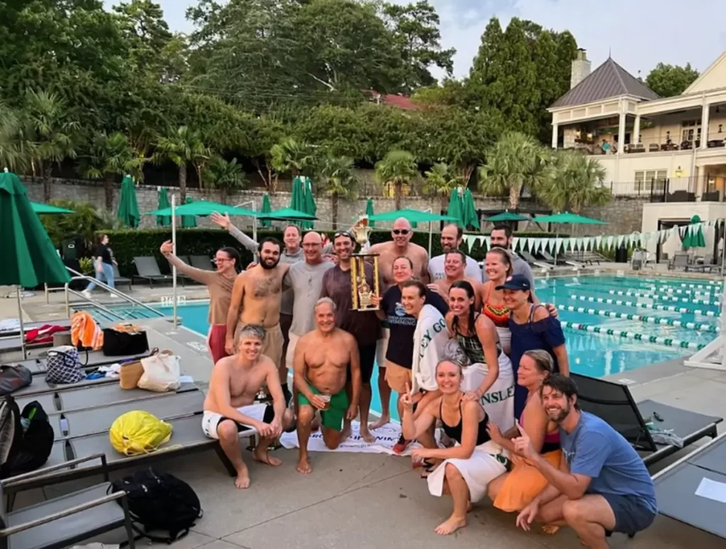 a group photo of about 15 swimmers of various ages by a pool with one holding a championship trophy